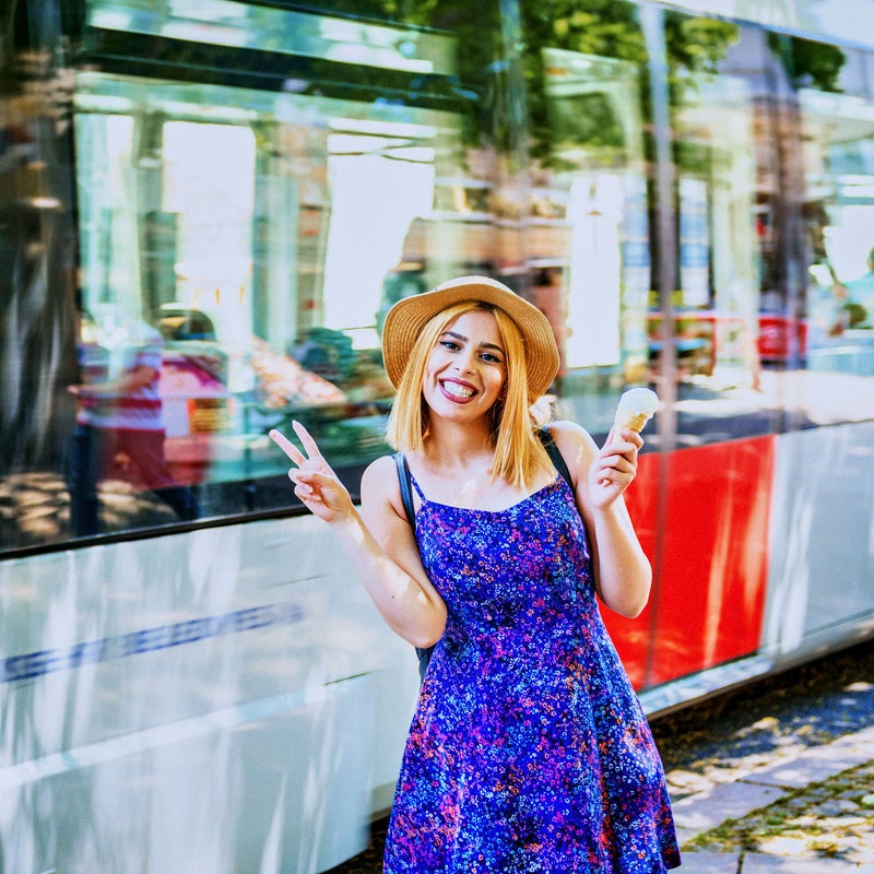 Mujer sonriente con vestido azul y bus detras, con un helado en una mano y haciendo la uve con la otra